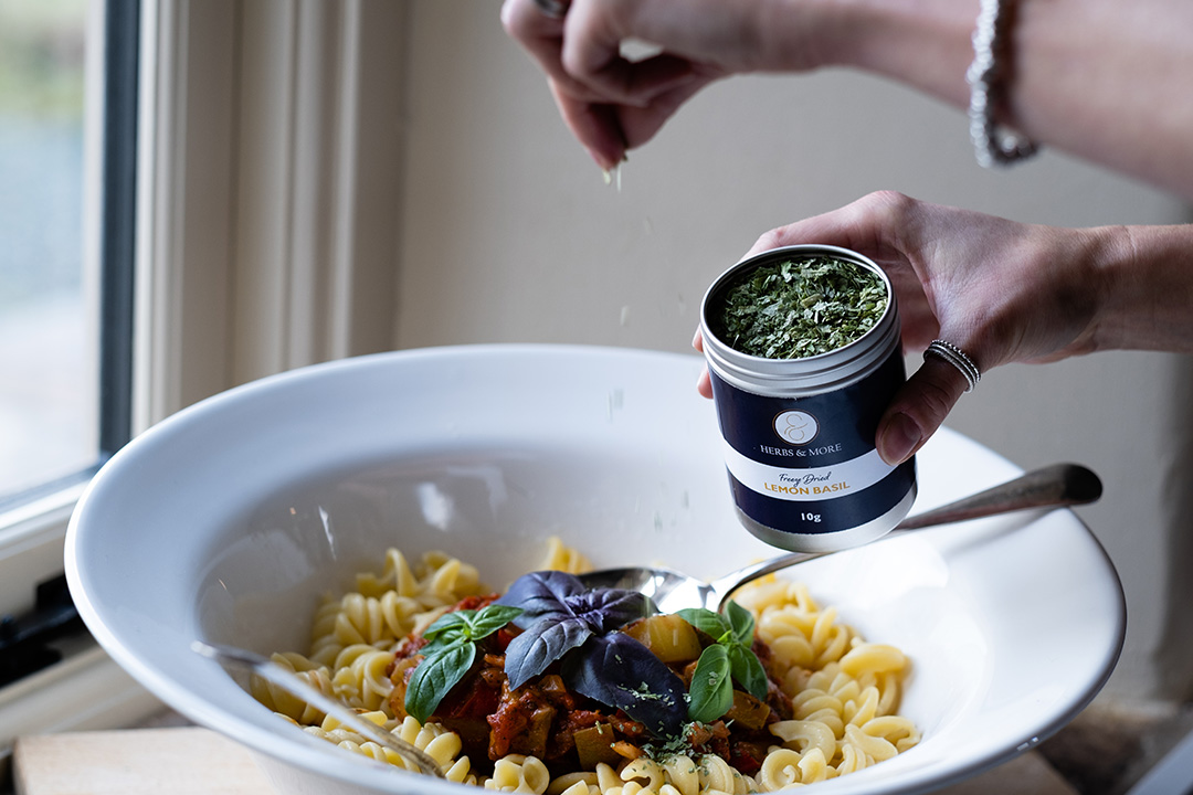 Tin of Lemon Basil freeze-dried herbs being sprinkled over pasta dish in white bowl